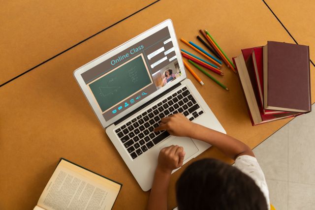 Child engaging in an online class using a laptop. Books and pencils on the desk suggest preparing notes or doing homework. Useful for topics related to virtual learning, distance education, technology in classrooms, homeschooling, or online study aids.