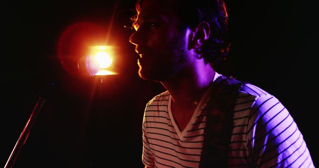 A young Caucasian male musician performs on stage, illuminated by dramatic stage lighting, with copy space. His silhouette against the bright backdrop creates an intimate atmosphere for the audience.