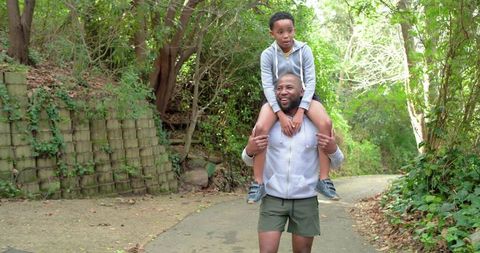 African american father carrying son on shoulders walking wooded trail for family bonding