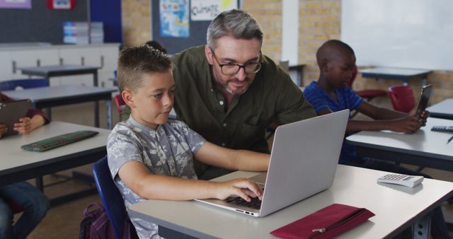 Diverse male teacher helping a schoolboy sitting in classroom using laptop. children in primary school.