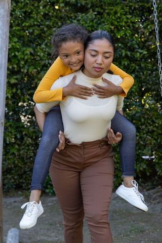 Mother carrying daughter piggyback in backyard by swing smiling family bonding moment