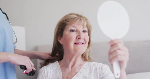 Senior woman checking hair in hand mirror while caregiver brushing hair in bright bedroom