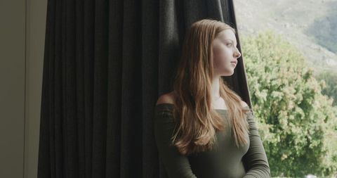 Young woman gazing out window, contemplative mood with natural sidelighting and greenery