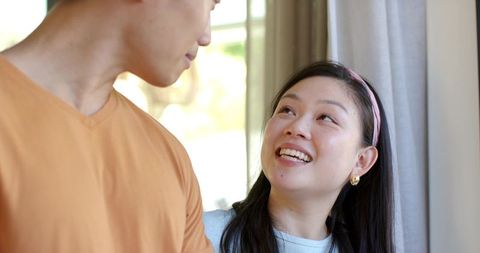 Asian couple sharing warm gaze by sunlit window woman wearing pink headband, smiling