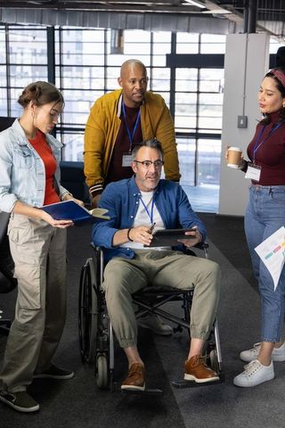 Diverse team collaborating with senior leader in wheelchair using tablet in modern office