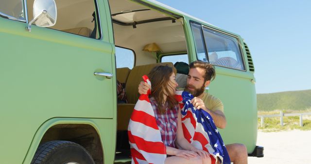 A young Caucasian couple shares a cozy moment inside a vintage van, with copy space. They are wrapped in an American flag, symbolizing a patriotic road trip or a Fourth of July celebration.