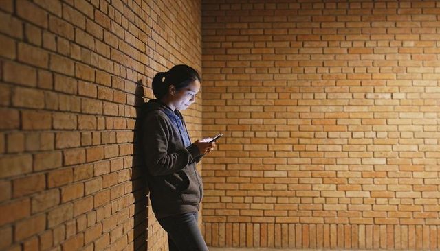 Young woman leaning on brick corner using smartphone in moody minimalist corridor