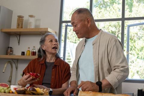 Senior asian couple preparing fresh fruit and slicing bread on sunlit modern kitchen