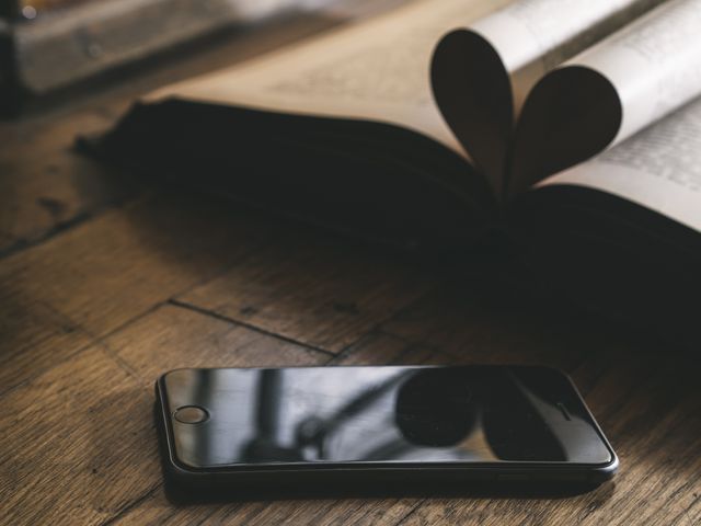 Smartphone lying on wooden table next to book with heart-shaped pages reflected on phone's screen. Great for themes on digital and literary connection, studying, educational purposes, romance in modern tech-driven world or valentines-related concepts.