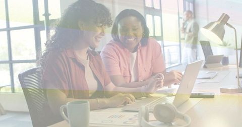 Two women collaborating at laptop reviewing charts and brainstorming in bright modern office