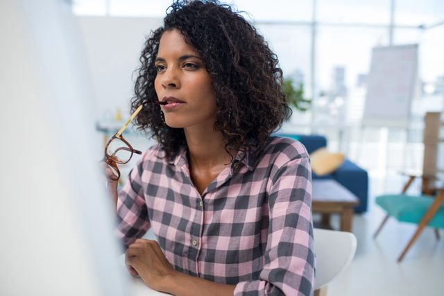 Female executive working on computer in modern office. She is wearing a plaid shirt and glasses, appearing focused and thoughtful. Ideal for use in business, technology, and productivity contexts, as well as articles on women in the workplace and modern office environments.
