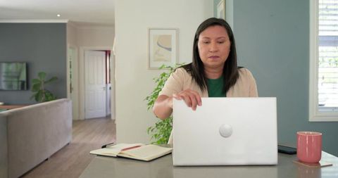 Mature woman working from home at kitchen island with laptop, notebook and pink mug