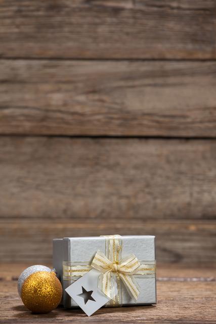 This image shows a wrapped gift box with a ribbon and tag, accompanied by gold and silver baubles on a wooden table. It is perfect for holiday-themed promotions, Christmas cards, festive blog posts, and seasonal advertisements.