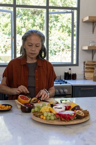 Senior woman preparing tropical fruit platter on marble kitchen counter with natural light