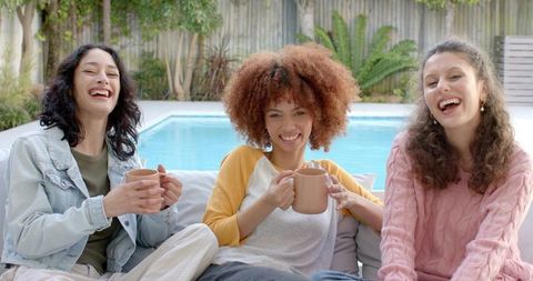 Multiracial friends laughing and sipping coffee on backyard sofa by pool