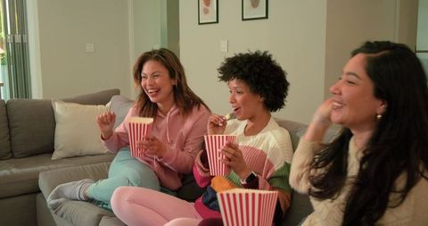 Multicultural friends laughing and eating popcorn on sectional couch during movie night