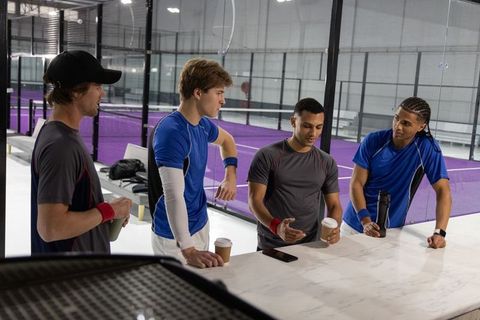 Multiracial team chatting over coffee by indoor padel courts, post-training break