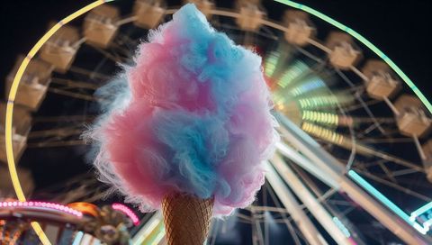 Towering pastel cotton candy on waffle cone with neon ferris wheel lights at carnival