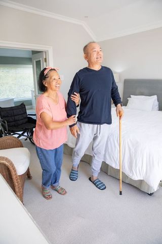 Asian senior couple standing in bright bedroom, man using cane while woman supporting