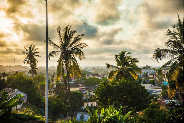 A serene early morning scene showing a coastal village with homes surrounded by lush greenery and palm trees under a vibrant sunrise sky. Ideal for travel blogs, nature enthusiasts, digital wallpapers, or promotional materials for serene vacation destinations.