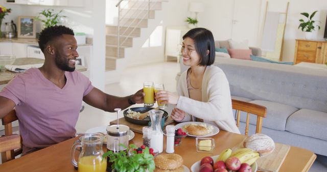 Couple enjoying a healthy breakfast together at a dining table in a cozy home. They are smiling and holding glasses of juice, indicating a pleasant morning together. Ideal for themes related to healthy living, relationships, hospitality, and home lifestyle. Suitable for use in advertisements, blogs, or articles focusing on healthy eating habits, couple activities, or morning routines.