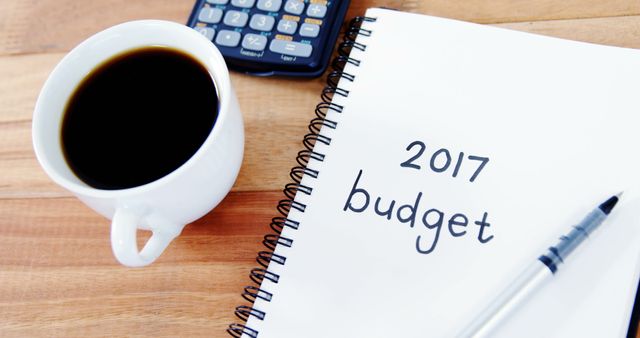 A cup of coffee sits next to a calculator and a notebook with 2017 budget written on it, with copy space. It suggests a setting of financial planning or budget review, in a business or personal finance context.