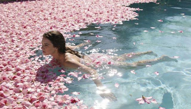 Floating woman in sunlit pool surrounded by pink flower petals for relaxation