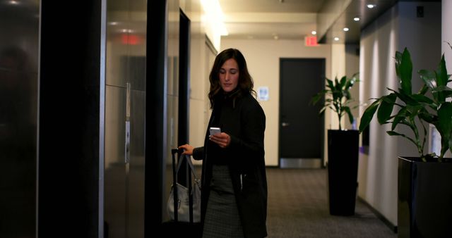 Businesswoman stands in a modern office corridor looking at her phone while holding a suitcase and a purse. The environment features sleek design with large planters and overhead lighting. Ideas for use include business travel, corporate communications, modern workplace, and professional work settings.