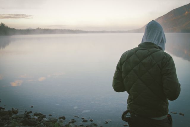 A person in a hoodie stands alone by the edge of a calm lake during dawn. The mist over the lake adds to the tranquil and serene atmosphere. This can be used in contexts of solitude, peaceful reflection, and connection with nature and for promoting outdoor and adventure activities or mental health themes.