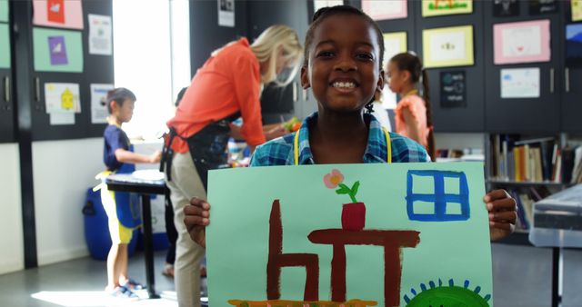 Smiling African American child displaying a colorful drawing in an engaging classroom setting with other students visible in the background working on their own projects. Perfect for articles or advertisements focusing on childhood creativity, art education, or celebrating artistic school activities.