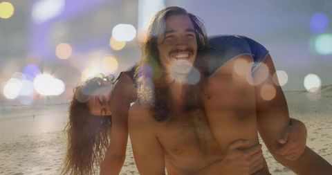 Carefree shirtless man carrying woman piggyback on sunny beach with bokeh light effects
