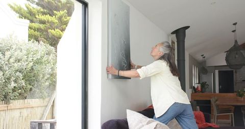 Senior woman balancing on sofa hanging artwork beside glass door in bright modern living room