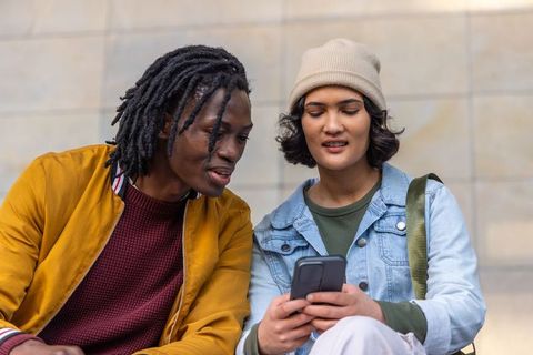 Diverse friends sharing smartphone on urban steps, wearing mustard jacket and denim jacket