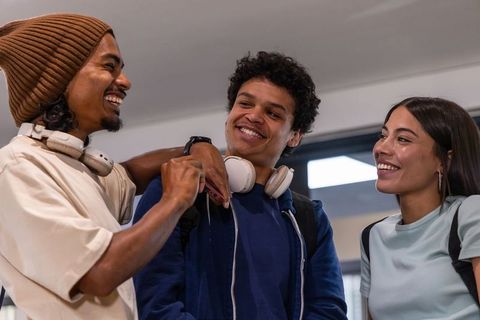 Multicultural teens laughing in campus hallway wearing headphones and backpacks