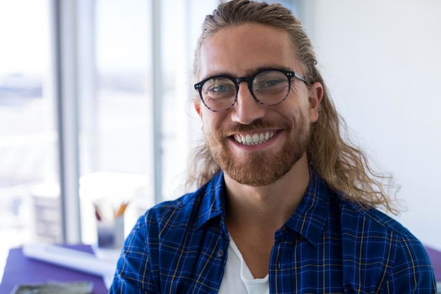 Young male architect with long hair and glasses smiling in a modern office. Ideal for use in articles or advertisements about architecture, creative professions, modern workspaces, and professional lifestyle.