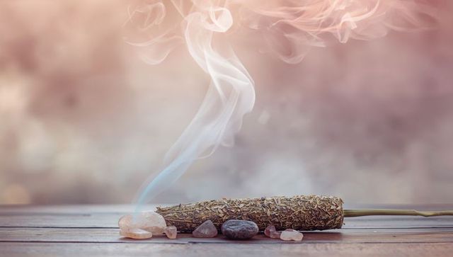 Smoldering sage bundle releasing delicate smoke with rose quartz and pebble on wooden plank