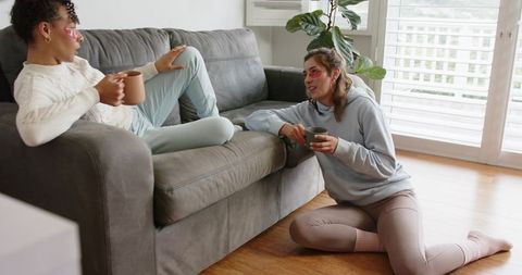 Cozy friends chatting on gray sofa wearing under-eye patches holding mugs in living room