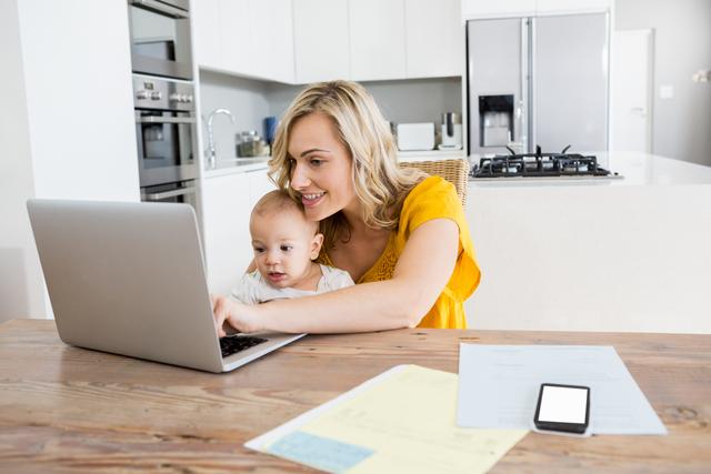 Mother using laptop with baby boy in kitchen at home