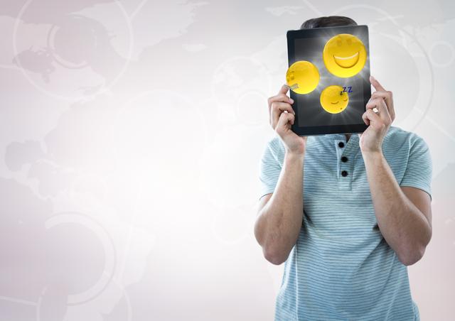 Man holding a tablet showing various smiling emojis in front of his face. The background features a world map and technological elements, adding a modern, digital feel. Useful for themes involving digital communication, expressions of emotions through technology, playful anonymity, and the intersection of technology and human interaction.