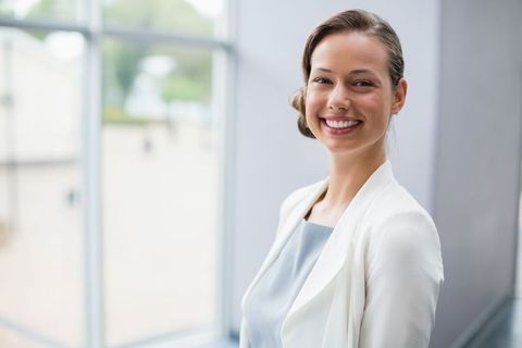 woman stands confidently near large window in modern office lobby, portraying a professional setting. her stylish white blazer and welcoming smile convey a sense of elegance and capability. ideal for corporate websites, business presentations, and marketing materials that highlight professional environments or personal success.