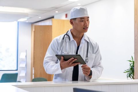 Asian male doctor using tablet at modern clinic reception, wearing lab coat and stethoscope