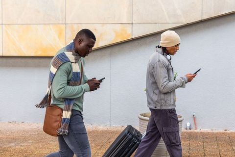 Two travelers walking using smartphones pulling rolling suitcase wearing casual jackets and beanie