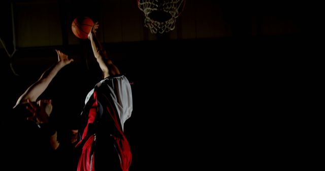 Basketball player in action, dunking the ball during an intense game under dim lighting. Ideal for use in sports-related articles, advertisements, and motivational content focusing on athleticism, teamwork, and competition.