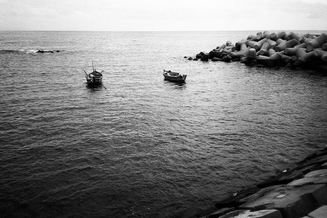 Two small boats moored near a rocky breakwater in the ocean. The boats and breakwater are seen in a wide view, with emphasis on the empty space of the calm water and smooth horizon under a cloudy sky, lending a minimalist and serene feel. Ideal for themes of tranquility, solitude, and coastal life.
