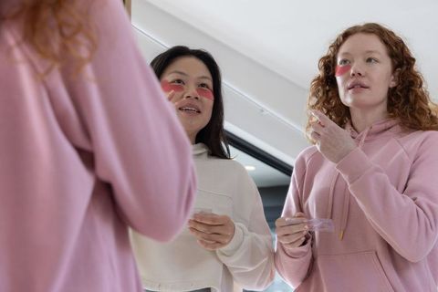 Friends applying pink under-eye gel patches at mirror for home skincare routine