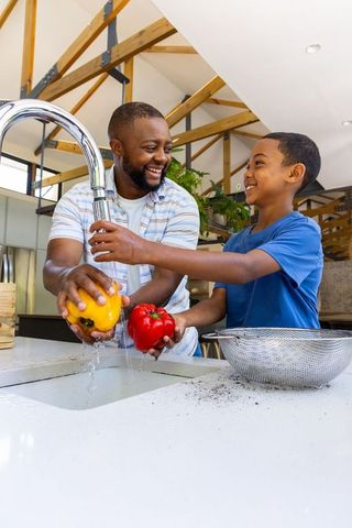 African american father and son smiling while washing bell peppers together at modern kitchen sink