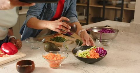 Diverse female friends seasoning colorful grain bowl with fresh vegetables in kitchen