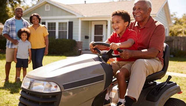 African american grandfather guiding grandson riding lawn tractor in sunny suburban yard