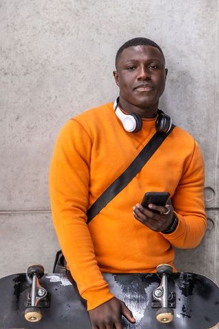 Urban skater standing with smartphone and headphones in orange crewneck against concrete