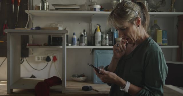 Woman standing in a workshop with shelves of tools and supplies in the background, using a smartphone intently. Great for illustrating themes of technology integration into everyday life, tech-savvy adults, focus and concentration, and casual, informal work environments.